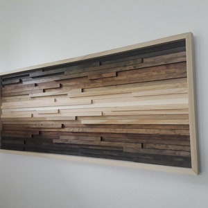 May include: A wood wall art piece with a gradient of brown, beige, and white wood strips arranged in a horizontal pattern. The wood strips are framed in a light wood frame.