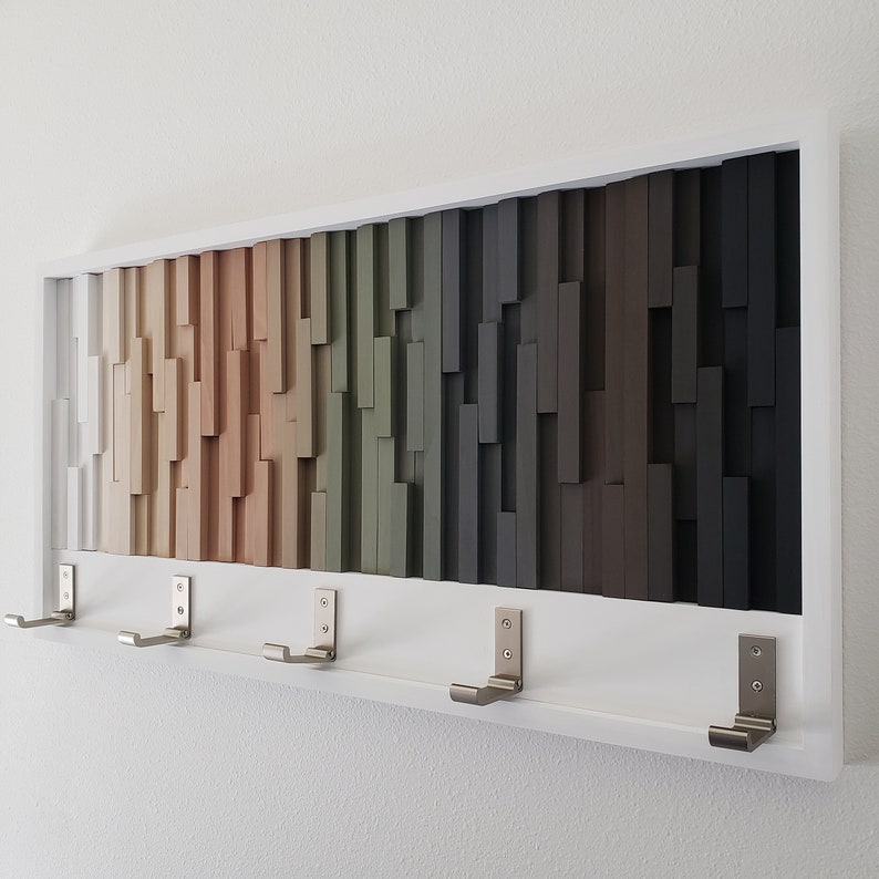 May include: A white wall-mounted coat rack with a wood slat design in shades of beige, green, and brown. The rack has five metal hooks.
