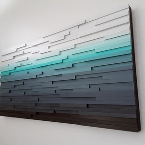 May include: A wall art piece made of wood, painted in shades of white, teal, and gray. The wood pieces are arranged in a staggered pattern to create a gradient effect.