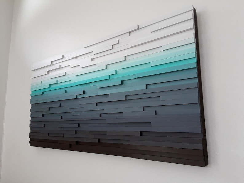 May include: A wall art piece made of wood, painted in shades of white, teal, and gray. The wood pieces are arranged in a staggered pattern to create a gradient effect.