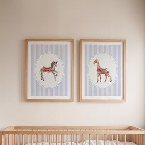 May include: Two framed prints with illustrations of a carousel horse and a giraffe. The artwork is set against a light blue and white striped background within a white oval. The frames are made of light-coloured wood.