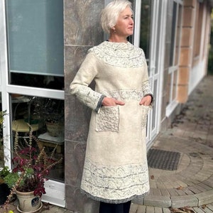May include: A long, white knitted sweater dress with a grey and white patterned trim. The dress has two large pockets on the front.