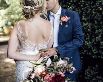 May include: A bride and groom kiss in a garden setting. The bride is wearing a white lace wedding dress with a low back and a floral crown. The groom is wearing a blue suit with a white shirt and a tie. The bride is holding a large bouquet of pink, red, and white flowers.