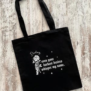 Könnte beinhalten: Schwarze Einkaufstasche mit weißem Text, der "Darling, even your darkest desires whisper my name." lautet. Der Text wird von einer Grafik einer skelettartigen Hand mit einer Rose begleitet.