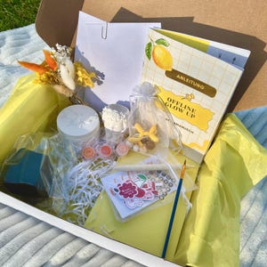 Könnte beinhalten: Eine offene Box voller Bastelmaterialien, darunter ein Buch mit einer Zitronenillustration und dem Text "OFFLINE GLOW UP". Die Box enthält einen weißen Behälter, kleine Farbtöpfe, Aufkleber, einen Pinsel und einen kleinen Strauß Trockenblumen.