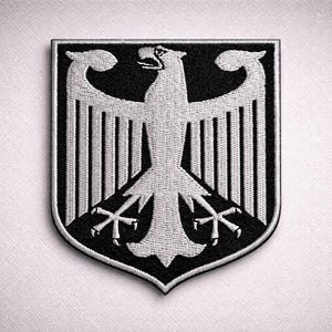 May include: Embroidered patch featuring the German coat of arms, a black shield with a white eagle emblem. The eagle has outstretched wings and is centered on the shield. The patch is a crest shape with a black border.