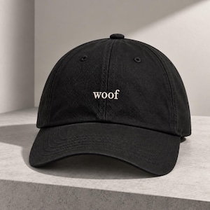 May include: A black baseball cap featuring the word woof embroidered in white lowercase serif letters on the front centre panel. The cap has a structured crown, a curved brim, and stitched eyelets for ventilation on each of the six panels.