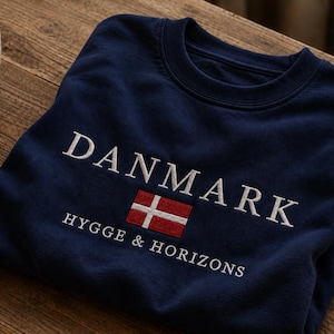 May include: A navy blue crewneck sweatshirt featuring white embroidered text that reads DANMARK and HYGGE & HORIZONS, with a small rectangular embroidered flag of Denmark in red and white positioned between the two lines of text.