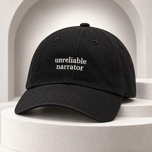 May include: A black baseball cap featuring the text "unreliable narrator" embroidered in white thread on the front panel. The cap has a structured crown, a curved brim, and stitched eyelets for ventilation, presenting a classic six-panel design.