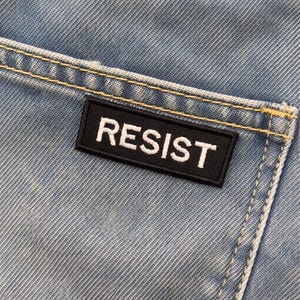 May include: Close-up of light blue denim fabric with a black rectangular patch. The patch has the word "RESIST" embroidered in white capital letters. Yellow stitching is visible along the seams of the denim.
