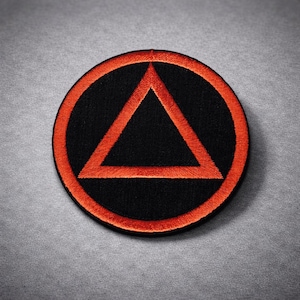 May include: A circular black patch with an orange embroidered border and a central orange triangle. The patch is likely an emblem or logo, suitable for application on clothing or accessories. The design is simple yet striking.