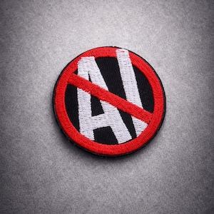 Pode incluir: Patch bordado com fundo preto, borda vermelha e as letras brancas "AI" riscadas com uma linha diagonal vermelha. O patch é circular e tem uma aparência costurada.