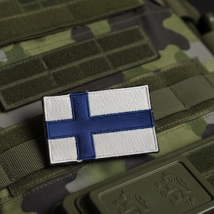 May include: A rectangular patch displaying the Finnish flag, a blue cross on a white field. The patch is attached to a camouflage tactical vest, with green straps and panels. The background has a camouflage pattern.