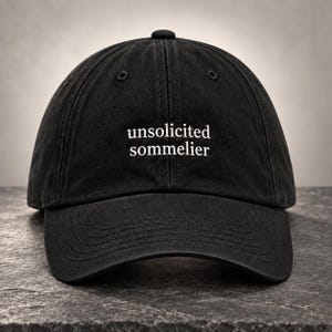 Op de afbeelding: Een zwarte baseballpet met de wit geborduurde tekst "unsolicited sommelier" in het midden op het voorpaneel. De pet heeft een gestructureerde kroon, een gebogen klep met zichtbare stiksels en ventilatiegaatjes op de bovenste panelen.