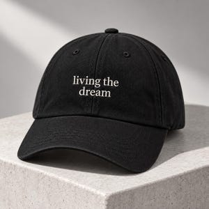 Könnte beinhalten: Eine schwarze Baseballkappe mit dem gestickten weißen Text "living the dream" mittig auf der Vorderseite. Die Kappe hat einen gebogenen Schirm, einen Knopf an der Oberseite und genähte Ösen zur Belüftung, gefertigt aus festem Baumwollstoff.