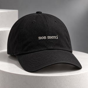 Op de afbeelding: Een zwarte baseballpet met de tekst "non merci" geborduurd in wit garen op het voorpaneel. De pet heeft een gestructureerde kroon, een gebogen klep met zichtbare stiksels en een knoopje bovenop in het midden van de bol.