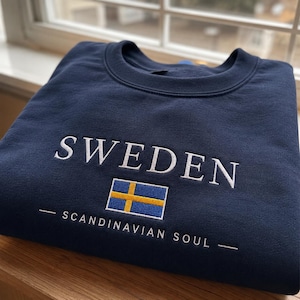 Könnte beinhalten: Ein marineblaues Sweatshirt mit Rundhalsausschnitt und weißem Sticktext, der SWEDEN lautet, über einer kleinen rechteckigen schwedischen Flagge in Blau und Gelb. Unter der Flagge steht der weiße Sticktext SCANDINAVIAN SOUL zwischen zwei Linien.