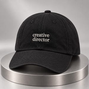 Puede incluir: Una gorra de béisbol negra con el texto "creative director" bordado en blanco centrado en el panel frontal. La gorra tiene una corona estructurada, una visera curva y ojales cosidos en los paneles superiores para ventilación y un diseño clásico.