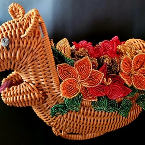 May include: A wicker squirrel figurine with a brown and orange beaded flower arrangement on its back. The squirrel is holding a small red ball in its paw.