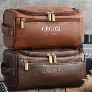 May include: Two brown leather toiletry bags with gold zippers and handles are stacked. The top bag is embossed with "GROOM" and the date "11.07.25". The bottom bag is embossed with the name "BARTON".