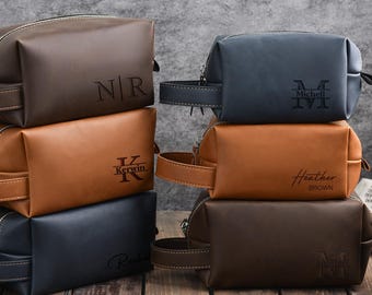 May include: A stack of personalized leather toiletry bags in various colors: brown, tan, and navy blue. Each bag features a zipper closure, a handle, and custom engraved initials or names. The bags are arranged on a wooden surface.