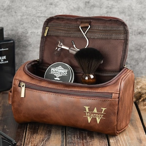 May include: Brown leather toiletry bag with a zipper closure, open to reveal a shaving brush, razor, and a tin of peppermint shaving soap. The bag is personalized with the name "Warner". A bottle of cologne and sunglasses are next to the bag.