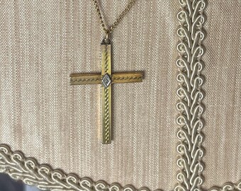 May include: A gold cross necklace with a small diamond in the centre. The cross is on a beige fabric background.