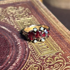 May include: A gold ring featuring a cluster of oval-cut, deep red gemstones, accented by smaller white stones. The ring is set on a detailed, antique-style book cover with gold embellishments, creating a vintage aesthetic.