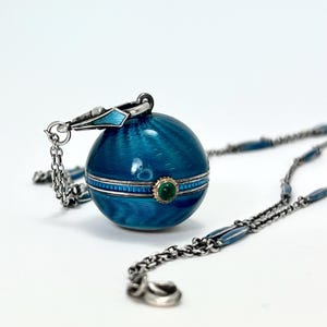 May include: A silver chain necklace with a blue enamel ball pendant. The ball has a silver band with a green gemstone set in the center.