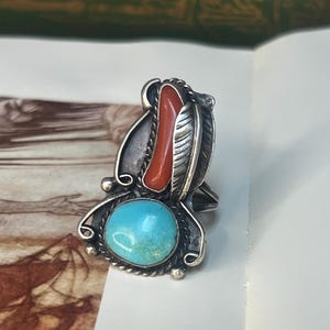 May include: A silver ring with a turquoise stone, a red coral accent, and a feather design. The ring has a detailed, ornate setting with a vintage aesthetic. The turquoise stone is round and a bright blue-green color.