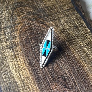May include: A silver ring with a long, diamond-shaped face. The face is inlaid with white, black, and turquoise-colored stones. The ring is set on a wooden surface.