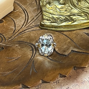 May include: A ring featuring a large, oval-cut, pale green gemstone set in a silver-toned metal frame. The setting includes small, clear accent stones and gold-toned metal details at the top and bottom of the central stone, creating a decorative border.
