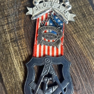 May include: A silver and black Masonic pin with a red, white, and blue ribbon. The pin features a compass and square with a hand holding a hammer and chisel. The ribbon has the text "J.R.O.U.A.M." and "OSANTIVILLE NO.141 COUNCIL".