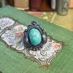 May include: A silver ring with an oval, turquoise stone. The ring has a detailed, ornate design with leaf and bead accents. The ring is set on a green surface with floral and text details.