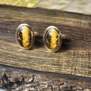 May include: A pair of gold-toned cufflinks with oval-shaped faces. Each cufflink features a carved cameo portrait in shades of brown and yellow. The cufflinks are set against a wooden surface.