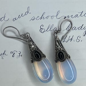 May include: A pair of silver-toned drop earrings. Each earring features a teardrop-shaped, opalescent stone with a black oval accent at the top. The earrings have a decorative, ornate design and are suspended from a simple hook.