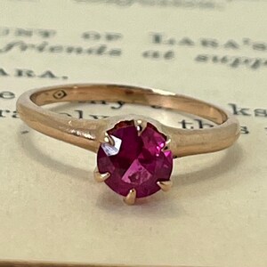 May include: A rose gold ring with a deep pink gemstone set in a prong setting.