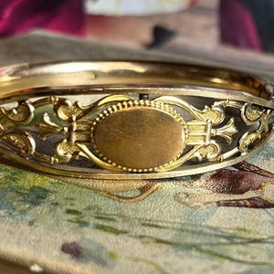 May include: A gold-toned bangle bracelet with an oval-shaped center piece. The bracelet has a delicate, intricate design with floral motifs.