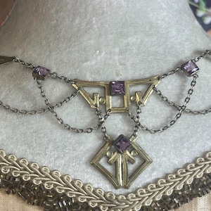 May include: A gold-tone necklace with a geometric design and purple stones. The necklace has a delicate chain and a pendant with a square shape.