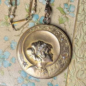 May include: A gold locket necklace with a detailed engraved design featuring a woman's profile and a crescent moon. The locket is adorned with small sparkling stones.