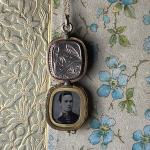 May include: A vintage gold-toned locket necklace with a rectangular pendant. The top section features an engraved floral design, while the bottom holds a small black and white portrait. The locket hangs on a delicate gold chain.