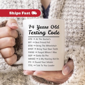 May include: White ceramic mug with the text "74 Years Old Texting Code" and a list of texting acronyms. The mug is held by a person. The mug is a standard 11 oz size.