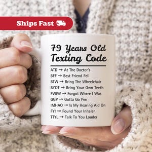 May include: White ceramic mug with the text "79 Years Old Texting Code" and a list of texting acronyms. The mug is held by a person wearing a gray sweater. The mug has a white handle.