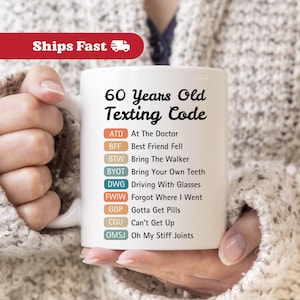 May include: White ceramic mug with the text "60 Years Old Texting Code" and a list of abbreviations with their meanings. The mug is held by a person wearing a sweater.