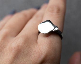 May include: A silver ring with an abstract, cloud-like design. The ring's band is dark, contrasting with the bright, reflective surface of the central shape. The ring is shown on a hand against a gray background.