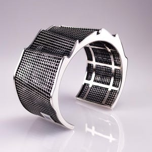 May include: A silver cuff bracelet with a geometric design. The bracelet features a mesh-like pattern in a dark gray color, contrasted by the polished silver frame. The design is modern and architectural.