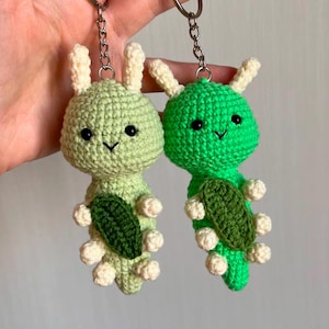 May include: Two crocheted green caterpillar keychains with white accents and black eyes. The caterpillars are holding green leaves.