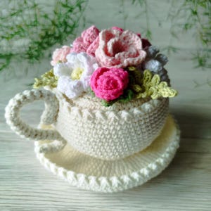 May include: A cream-coloured crocheted teacup and saucer set, adorned with a bouquet of handmade flowers in pink, white, and green hues. The teacup features a handle and rests on a matching saucer, creating a charming, handcrafted decorative item.