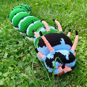 May include: A large, crocheted caterpillar toy in a grassy setting. The caterpillar features segments of green, light blue, and white, with a black head and pink and black legs. The toy is designed to resemble a caterpillar.