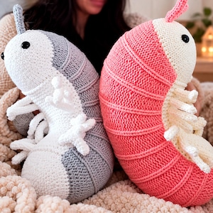 May include: Two plush toys shaped like isopods, one in grey and white and the other in pink and white. Each creature features a segmented body, multiple small white legs, a single antenna on top, and round black eyes made of plastic buttons.